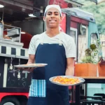 ovem cozinheiro sorridente com avental azul segurando dois pratos de pizza em frente ao seu food truck vermelho e preto, pronto para servir os clientes.