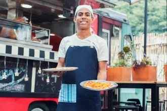 ovem cozinheiro sorridente com avental azul segurando dois pratos de pizza em frente ao seu food truck vermelho e preto, pronto para servir os clientes.