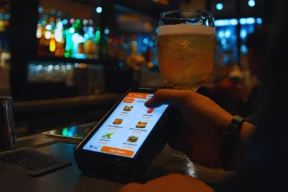 Pessoa toca a tela de vendas de uma maquininha no balcão de um bar, com um copo de cerveja ao fundo.