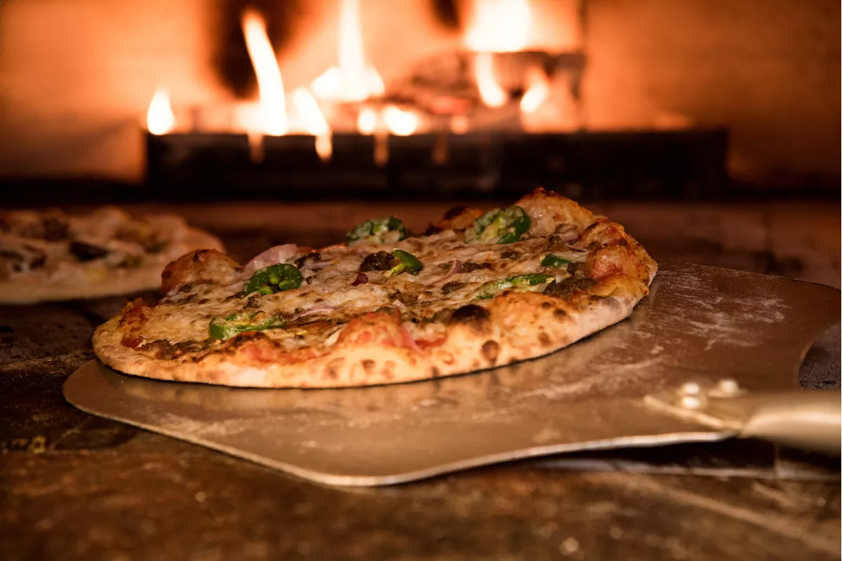 Pizza recém-saída do forno à lenha, sobre pá de madeira, com borda crocante e queijo derretido, representando o Dia da Pizza em restaurantes e delivery.

