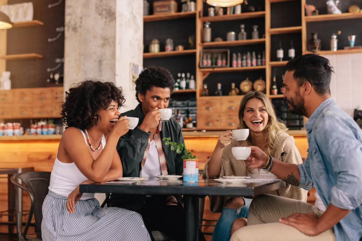 Grupo de quatro amigos sorrindo e tomando café em mesa de bar ou cafeteria, em clima descontraído e alegre, representando o Dia do Amigo em bares e restaurantes.

