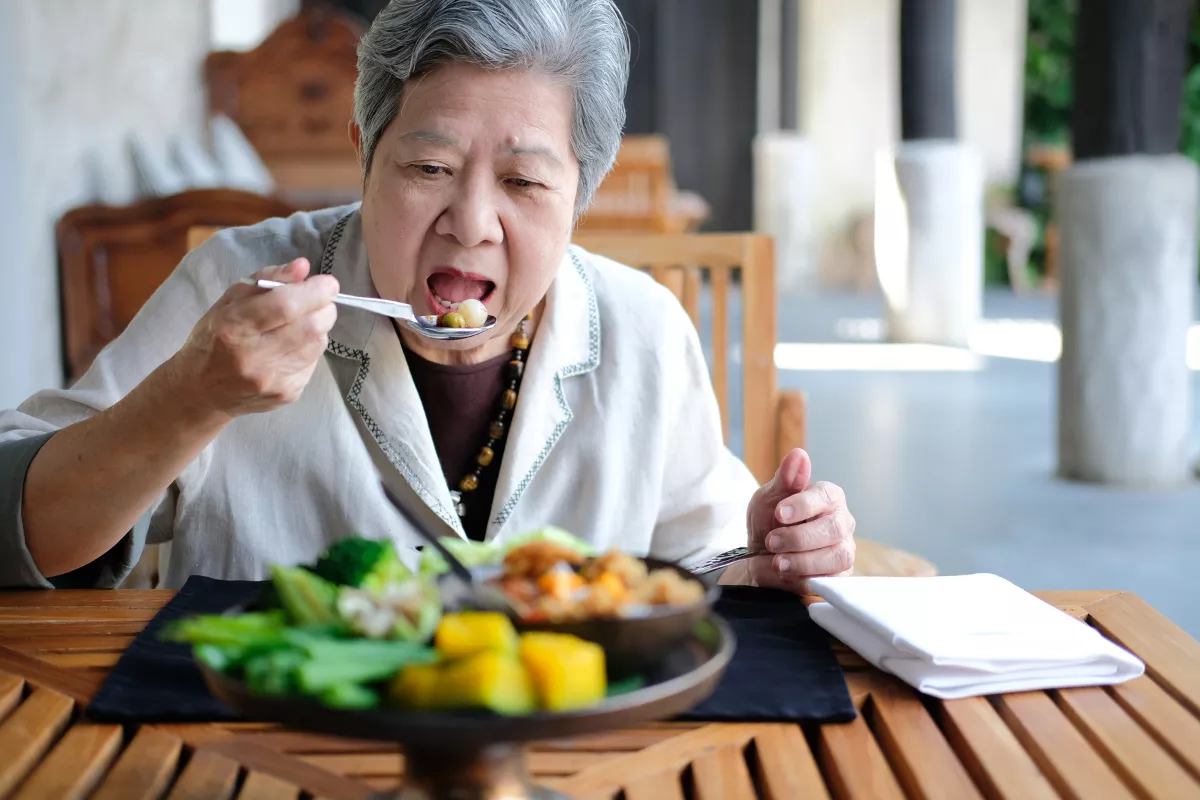 Senhora idosa apreciando refeição saudável em restaurante, com legumes e alimentos frescos no prato, simbolizando o almoço em família no Dia dos Avós.