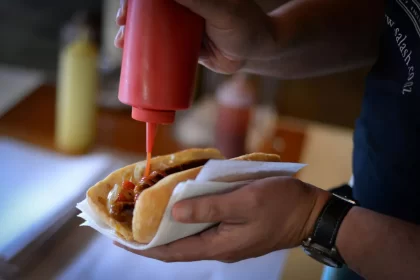 Pessoa aplicando ketchup em hot dog artesanal com cebola e molho barbecue, segurando o lanche com guardanapo, em ambiente informal de lanchonete.