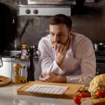Cozinheiro pensativo em cozinha de restaurante, com alimentos frescos na bancada, simbolizando os desafios da jornada noturna no food service.