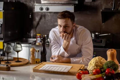 Cozinheiro pensativo em cozinha de restaurante, com alimentos frescos na bancada, simbolizando os desafios da jornada noturna no food service.