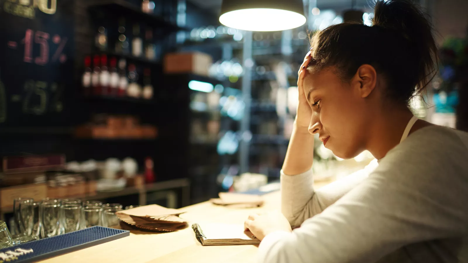 Funcionária de avental com a mão na testa, preocupada, apoiada no balcão de um bar à noite diante de um caderno aberto, com copos e garrafas ao fundo