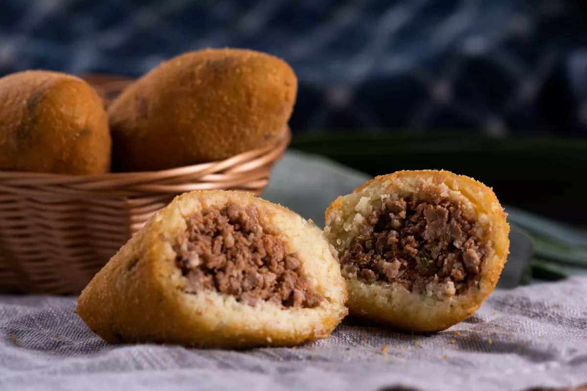 Bolinho de aipim crocante por fora e recheado com carne desfiada temperada, servido em cesto de palha, típico das festas juninas.