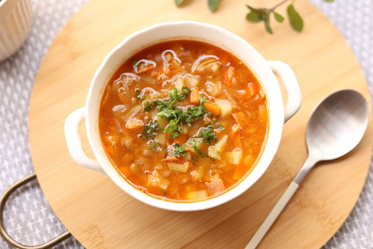 Cumbuca branca com sopa de legumes colorida, decorada com salsinha fresca, acompanhada de colher sobre tábua de madeira, típica de cardápio de inverno em restaurante.