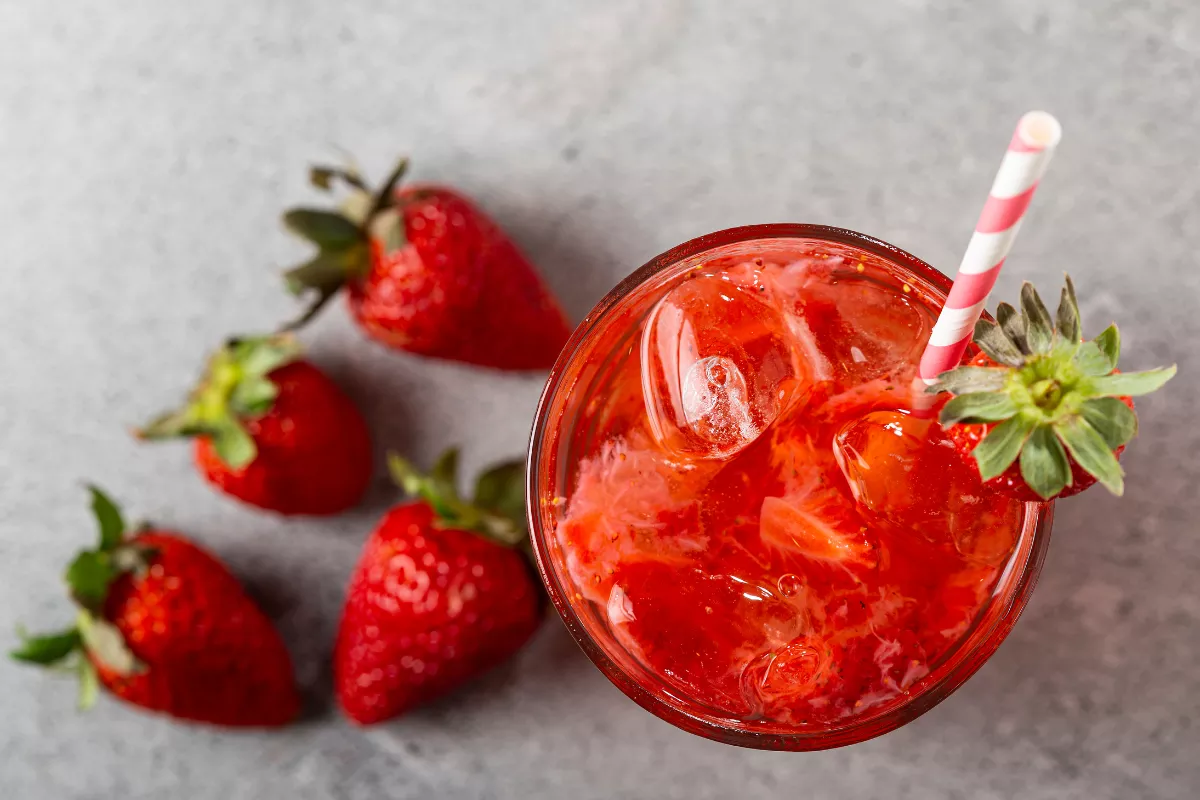 Drink refrescante à base de morango com gelo e canudo listrado, cercado por morangos frescos — sugestão de coquetel romântico para cardápios do Dia dos Namorados.

