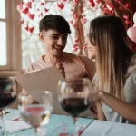 Casal sorridente sentado em restaurante decorado com corações vermelhos, analisando juntos um cardápio — clima romântico e descontraído para celebrar o Dia dos Namorados.
