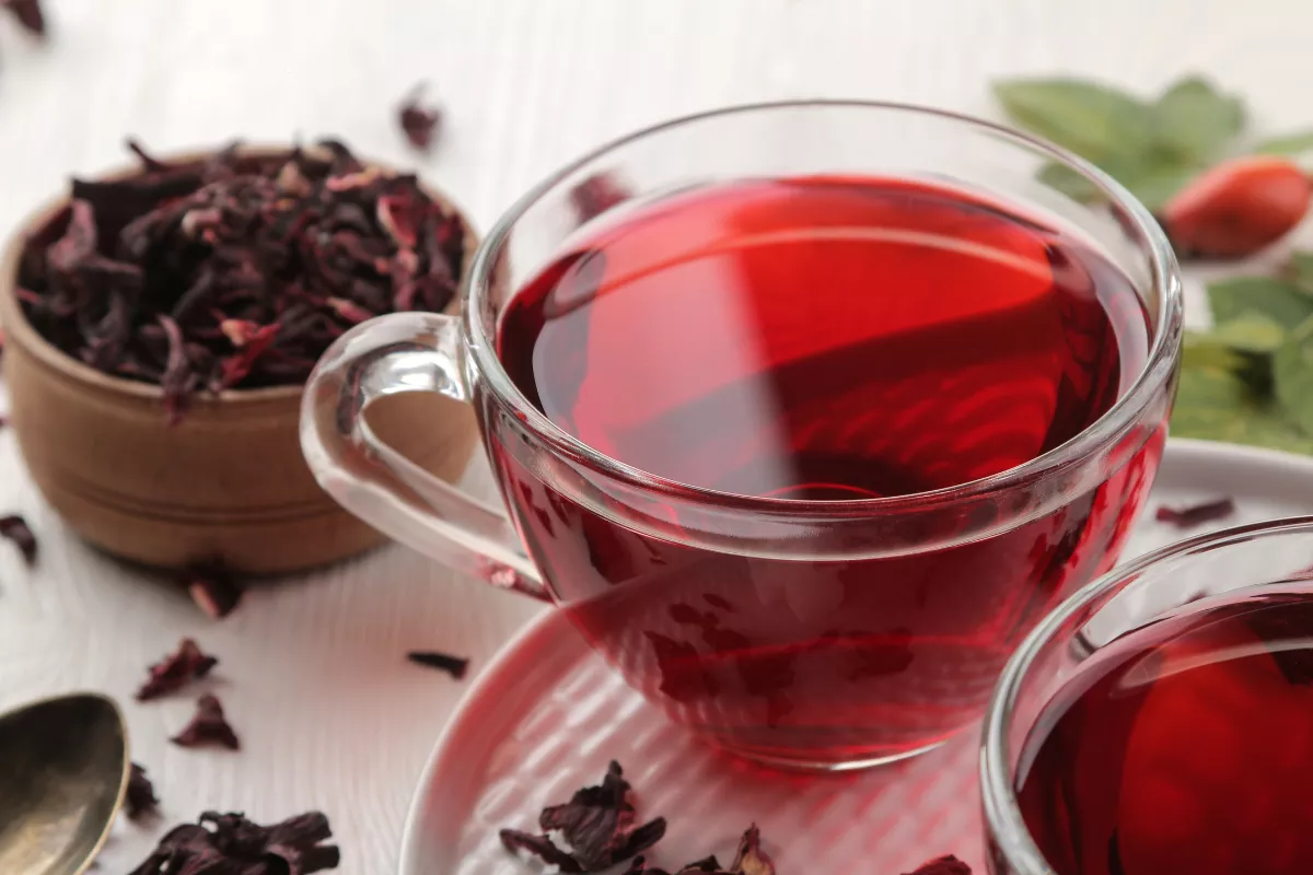  Xícara de chá de hibisco com líquido vermelho intenso, cercada por flores secas de hibisco e folhas verdes, bebida quente para complementar o cardápio de inverno.