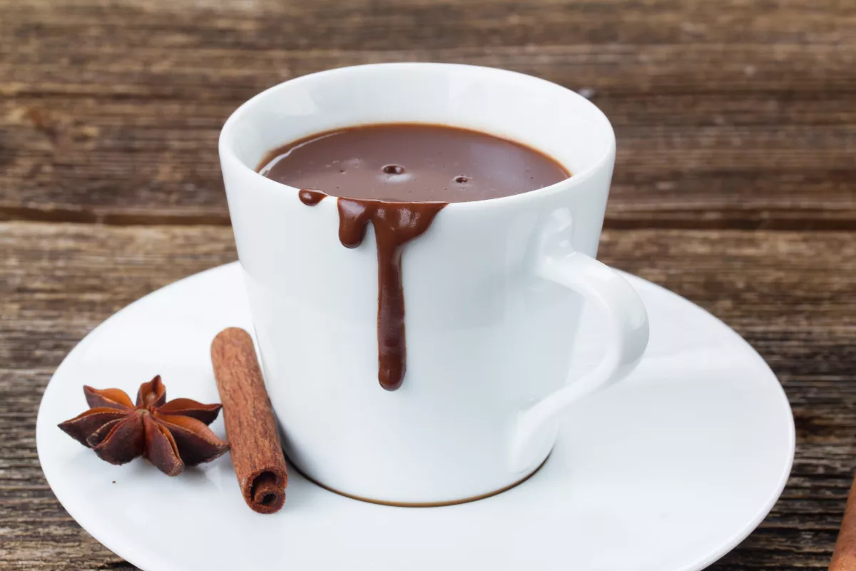 Xícara branca com chocolate quente cremoso escorrendo na borda, ao lado de canela em pau e anis-estrelado, sobre pires branco.