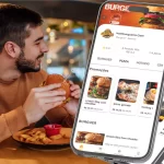 Homem sorridente em restaurante segurando hambúrguer com batatas fritas no prato, enquanto na frente aparecem celulares exibindo o cardápio digital de uma hamburgueria no sistema CNM, com promoções e opções de pedido online.