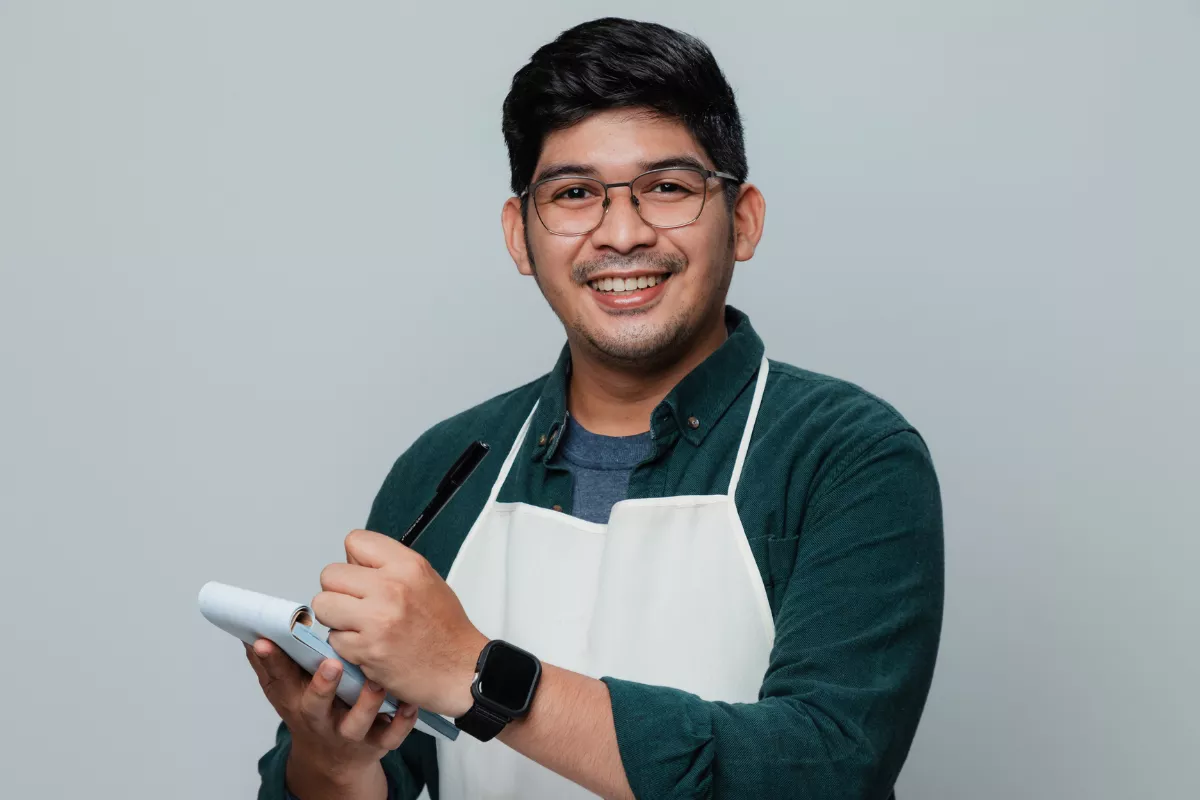 Garçom sorridente com avental, segurando bloco de anotações e caneta, representando trabalhadores de bares que atuam no período noturno.


