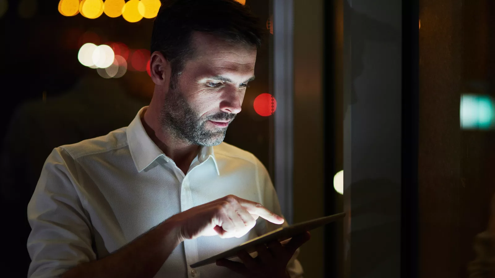 Gestor de restaurante de camisa branca consultando um tablet à noite, com o rosto iluminado pela tela e luzes desfocadas ao fundo, acompanhando os custos do turno noturno