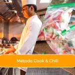 Chef profissional cozinhando em cozinha industrial com chama alta ao lado de alimentos preparados embalados a vácuo, representando o processo cook & chill em restaurantes.