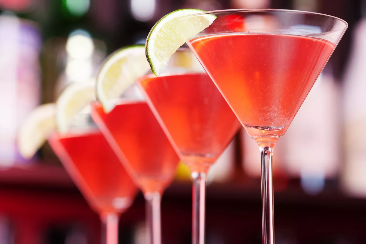 Fila de taças de martini com Cosmopolitans vibrantes, decoradas com fatia de limão. Bebidas coloridas e elegantes em ambiente de bar sofisticado.