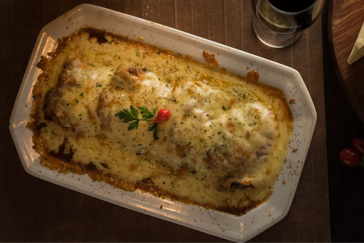 Escondidinho de carne seca gratinado com queijo, servido em travessa branca, polvilhado com ervas e decorado com pimenta e salsinha.