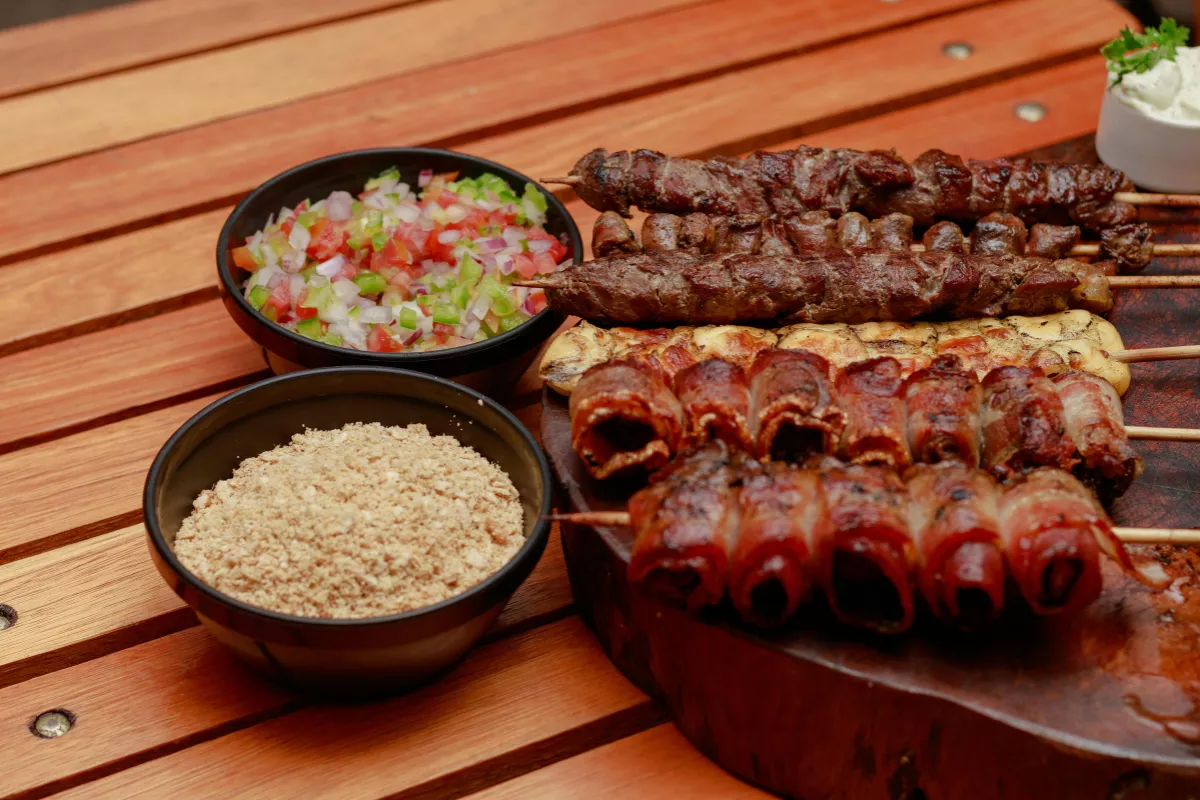Espetinhos variados com carne, frango e bacon servidos em tábua de madeira, acompanhados de vinagrete e farofa em potes, prontos para festa junina.