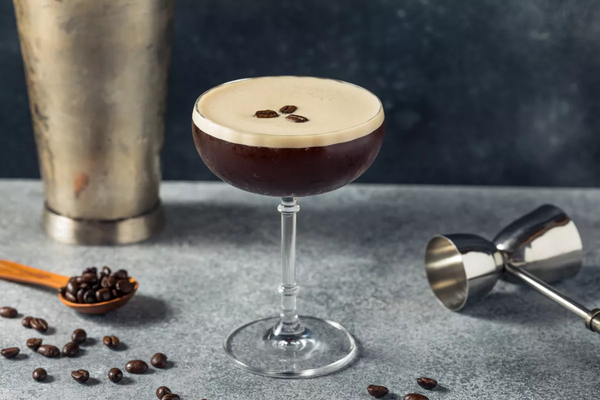 Taça coupe com um Expresso Martini sobre uma bancada de pedra, decorado com três grãos de café. Ao fundo, utensílios de bar e grãos soltos ajudam a compor o visual sofisticado do drink.