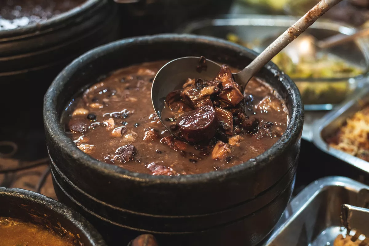 Panelas de barro com feijoada fumegante, com concha servindo feijão e carnes, prato brasileiro tradicional nos almoços de inverno em restaurante.