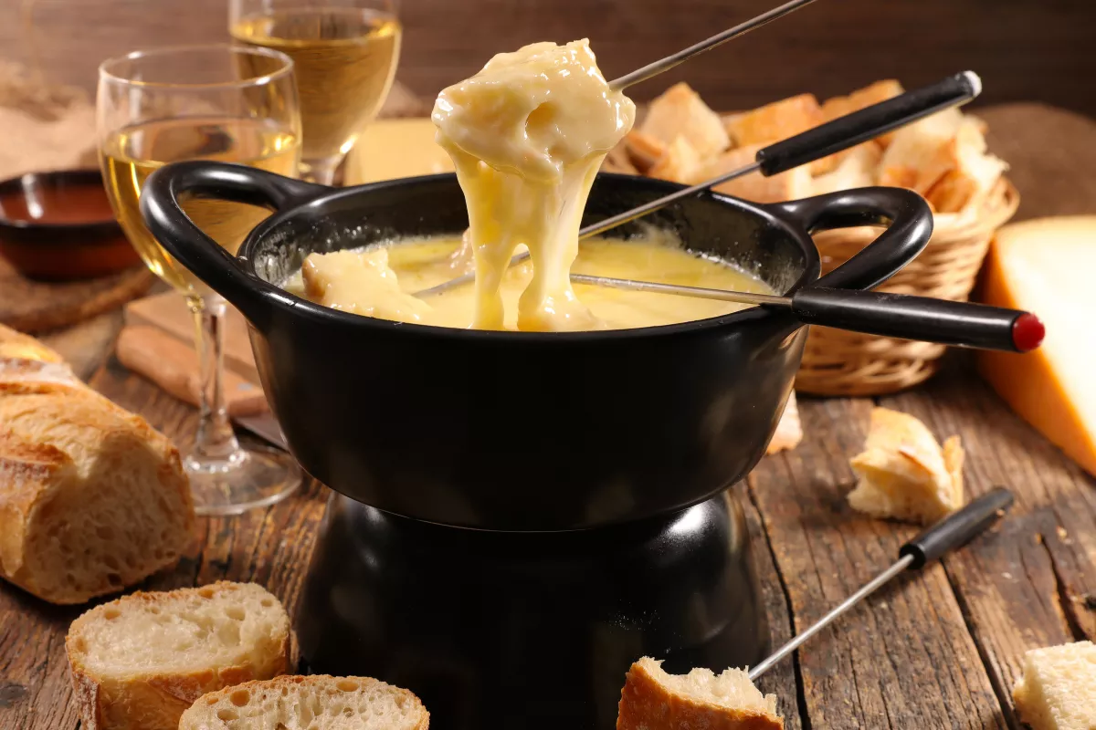 Fondue de queijo derretido servido em panela preta com garfos, pedaços de pão rústico e taças de vinho branco ao fundo, prato típico do inverno em restaurante.