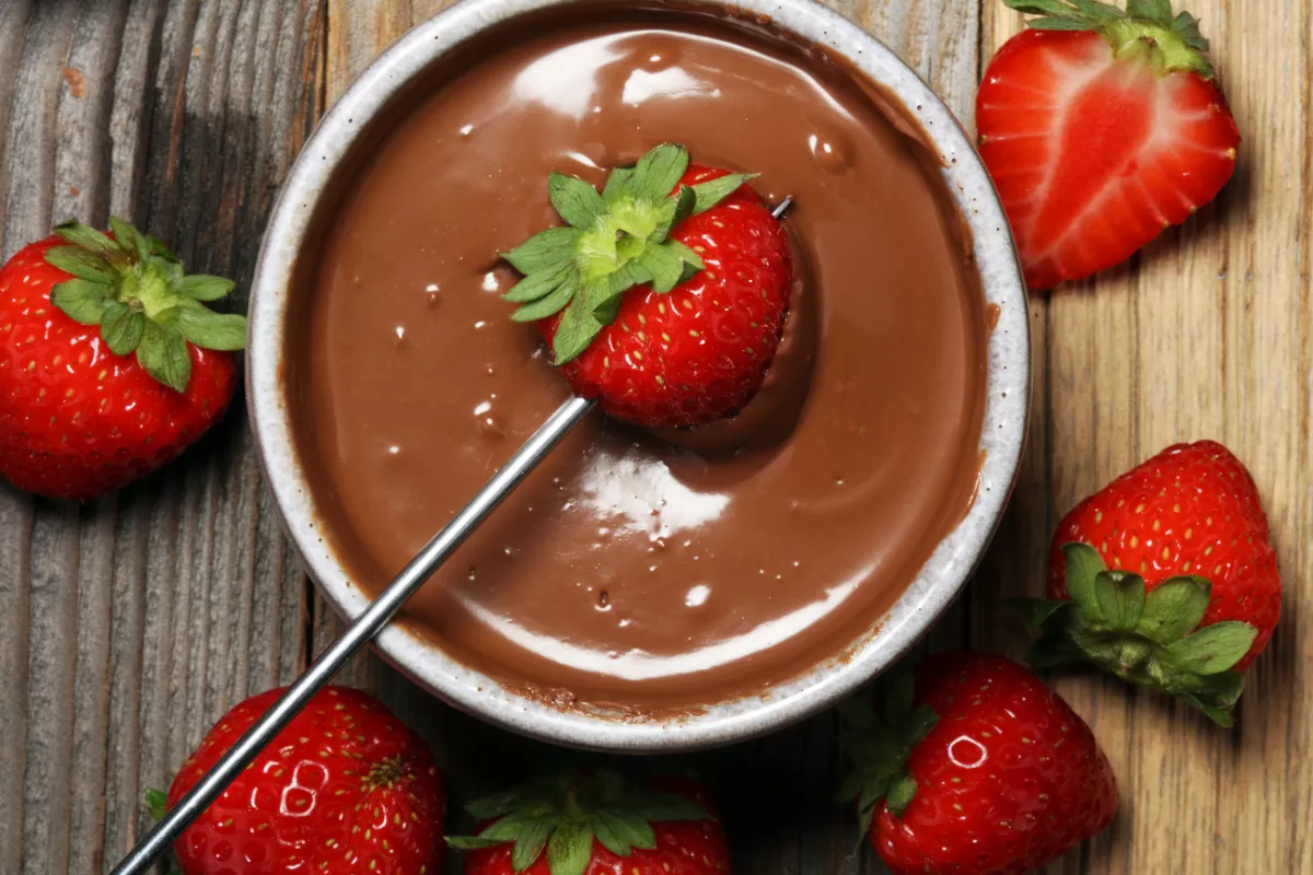Morango fresco sendo mergulhado em fondue de chocolate cremoso, com mais frutas ao redor — sobremesa clássica e romântica para jantares no Dia dos Namorados.