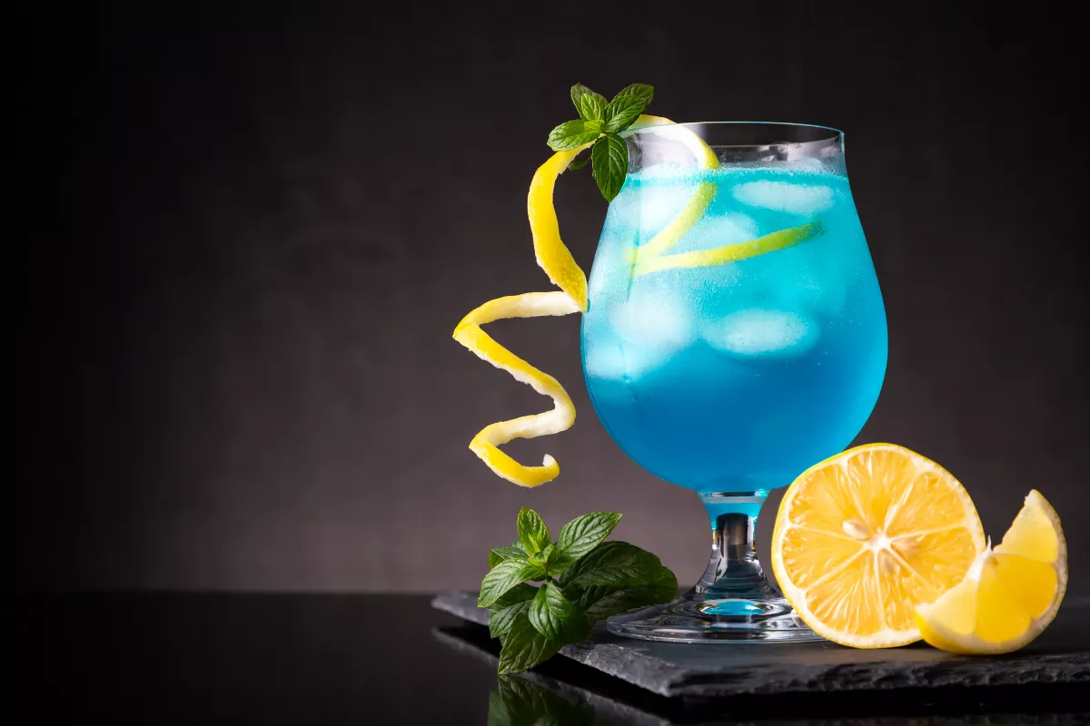 Taça arredondada com drink Blue Lagoon decorado com espiral de casca de limão, folhas de hortelã e limão siciliano ao lado. Drink visualmente impactante e refrescante.