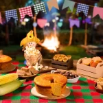 Mesa decorada com toalha xadrez e pratos típicos juninos como bolo de milho, milho cozido, pé de moleque, pipoca, paçoca e outras delícias ao ar livre, com bandeirinhas e fogueira ao fundo.