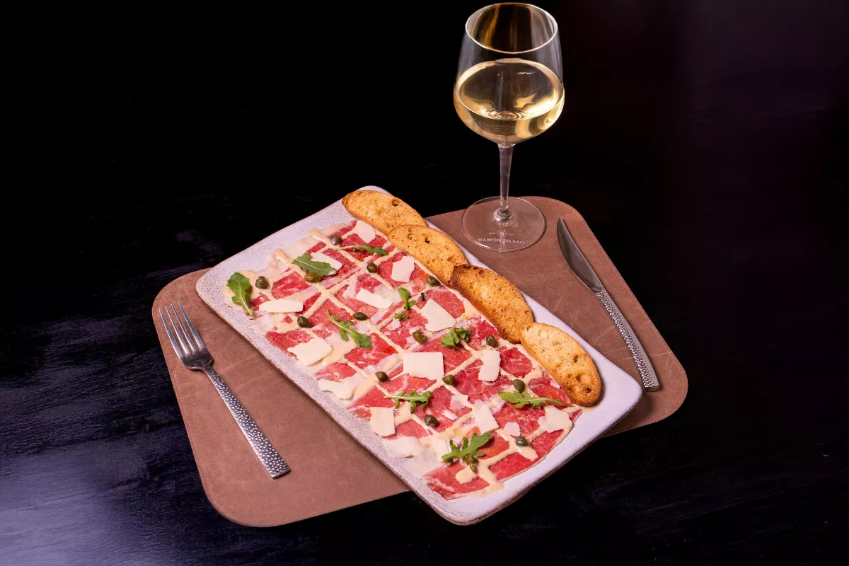 Carpaccio de carne bovina com lascas de parmesão, alcaparras e rúcula servido com torradas, acompanhado de taça de vinho branco — entrada elegante para menus fechados.