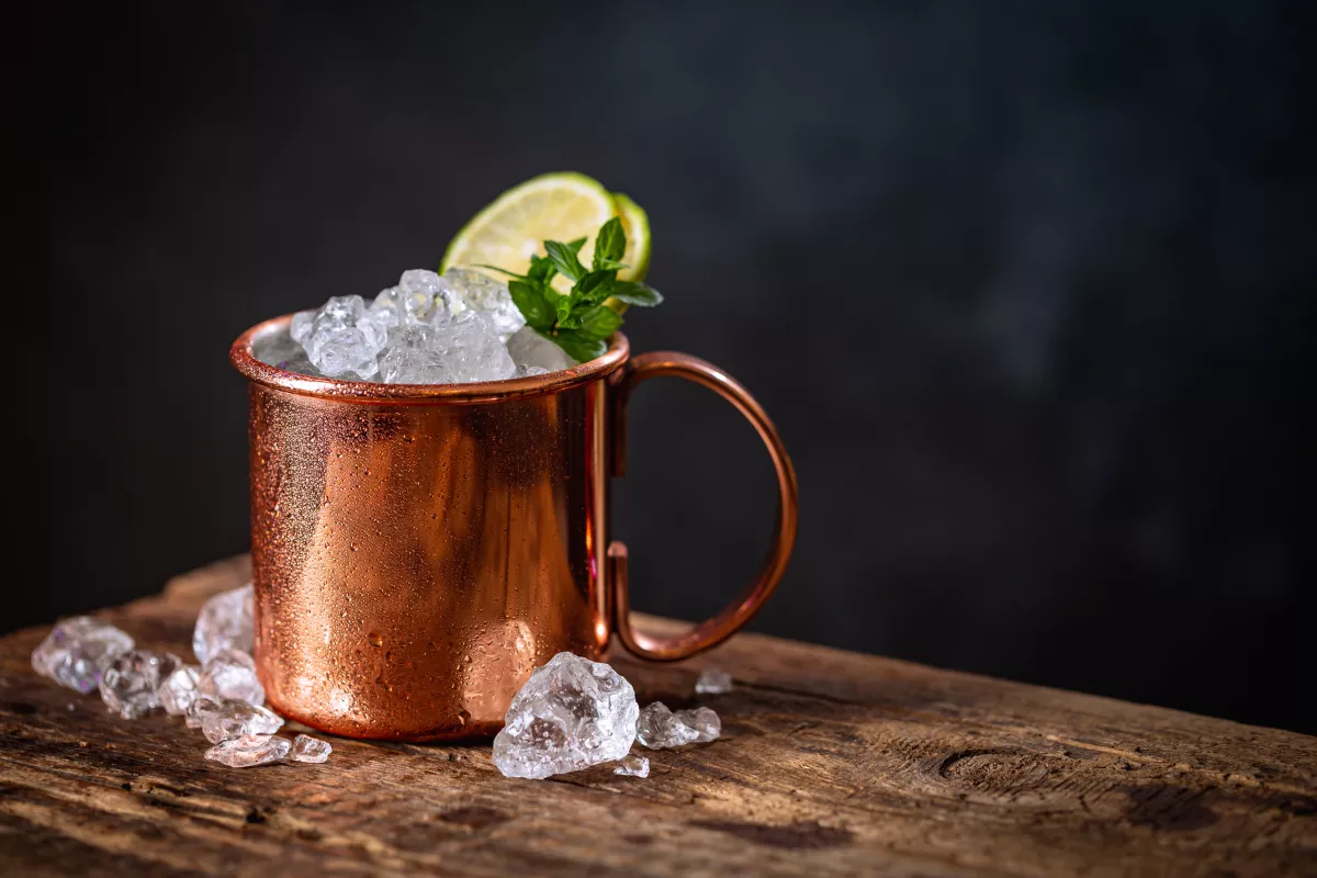 Drink Moscow Mule servido em caneca de cobre com muito gelo, rodela de limão e hortelã, sobre mesa rústica. Visual clássico e refrescante.