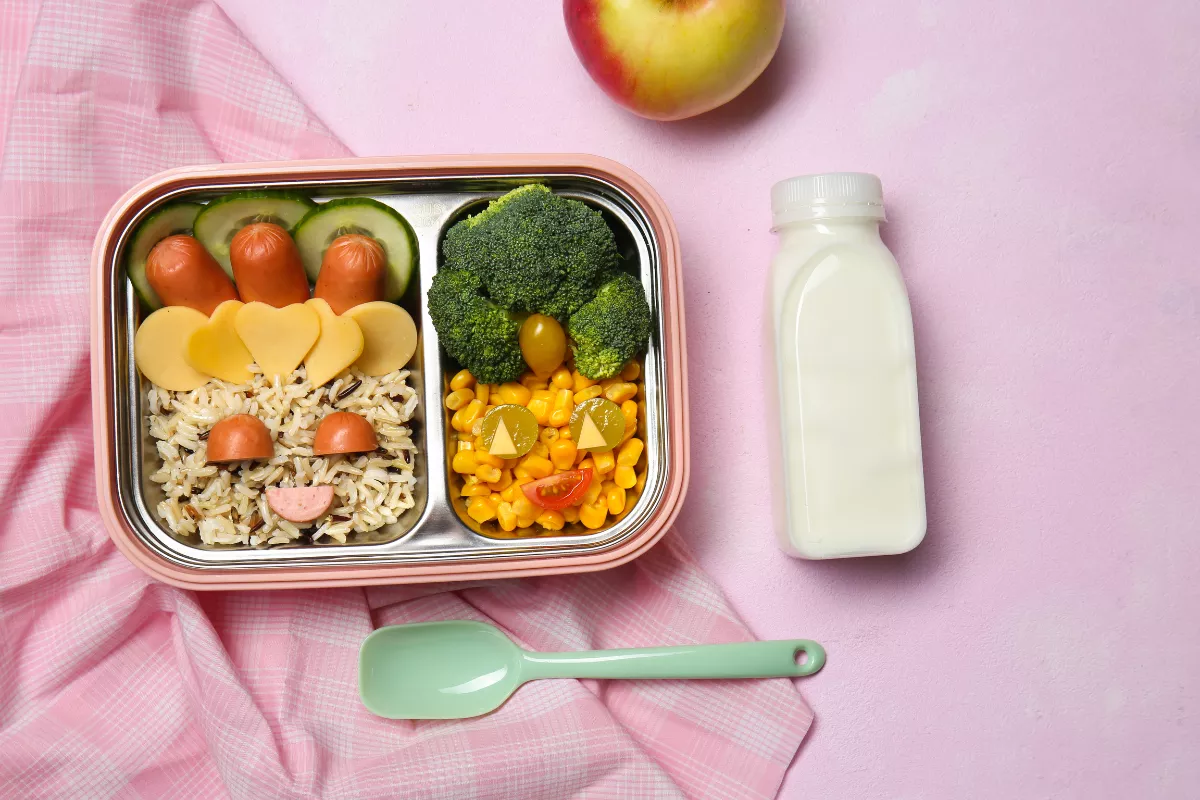 Marmita divertida e infantil com arroz em formato de rosto sorridente, salsichas, brócolis, milho e tomate cereja em uma lancheira rosa, acompanhada de suco branco e maçã sobre fundo rosa.