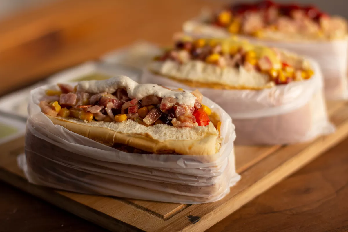 Hot dog no estilo brasileiro, servido em pão macio e envolto em plástico, recheado com milho, bacon, vinagrete e molho branco, sobre tábua de madeira.