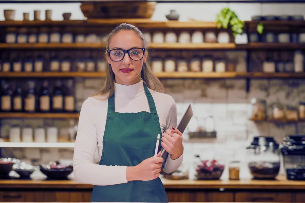 Funcionária de açaiteria com avental verde e óculos de grau, em frente a prateleiras de madeira repletas de potes de ingredientes, em ambiente moderno e acolhedor. A imagem transmite profissionalismo e organização, reforçando o conceito de gestão eficiente em açaiterias.