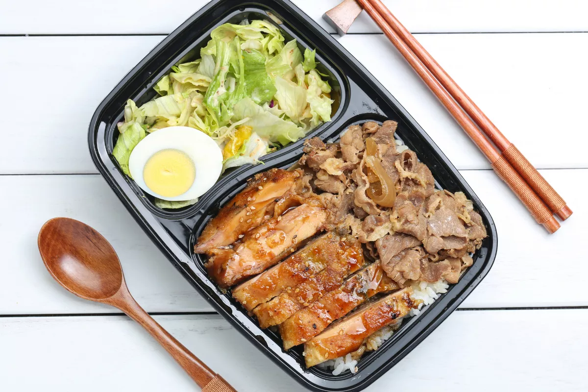 Marmita caseira em embalagem preta com arroz coberto por frango teriyaki, carne acebolada, salada de alface e ovo cozido fatiado, acompanhada de hashis e colher de madeira. 