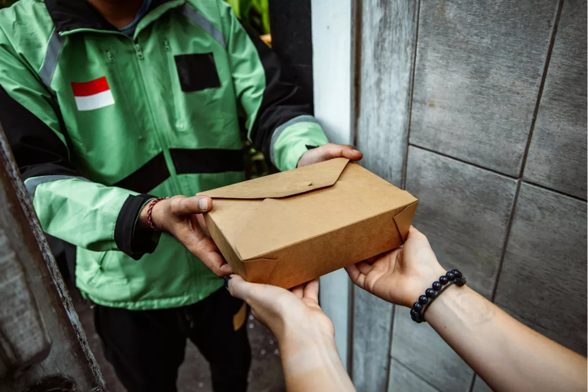Entrega de marmita feita por motoboy uniformizado, segurando embalagem de papel kraft sendo recebida pelas mãos do cliente em ambiente residencial. 