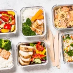 Marmitas gourmet com pratos elaborados como frango envolto em bacon, batatas rústicas, saladas frescas e risotos coloridos em embalagens de alumínio bem montadas.