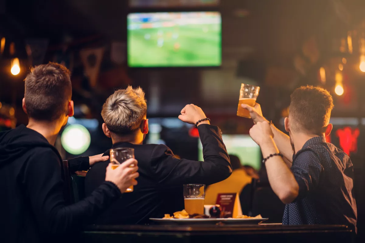 Três amigos assistindo a um jogo de futebol em bar, segurando copos de chope e comemorando animados enquanto acompanham a partida no telão.