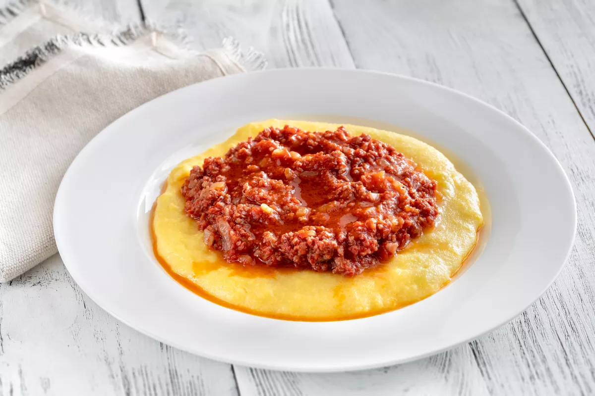 Prato de polenta cremosa coberta com ragu de carne moída ao molho vermelho, apresentado em prato branco sobre mesa rústica, opção reconfortante no frio.