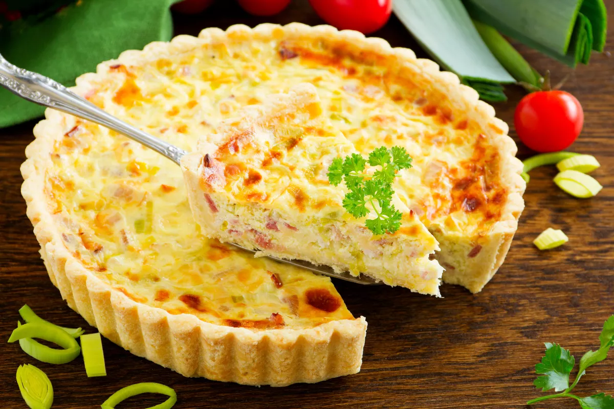 Quiche dourada e cremosa com pedaço sendo servido, decorada com salsinha, ao lado de tomates e alho-poró frescos, prato leve para lanches e jantares no inverno.