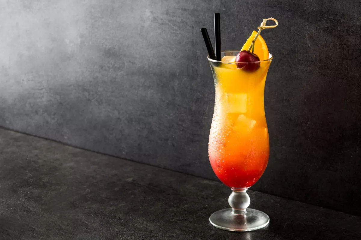 Drink Sex on the Beach servido em taça hurricane com degradê vibrante de laranja e vermelho, decorado com cereja, fatia de laranja e canudo. Bebida tropical com apelo visual forte.