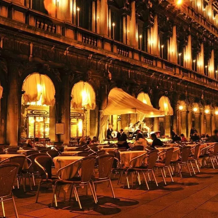  Fachada iluminada do Caffè Florian à noite, na Piazza San Marco, em Veneza. As mesas ao ar livre estão dispostas sob arcadas históricas, com cortinas brancas esvoaçantes e uma pequena orquestra tocando ao vivo, criando uma atmosfera elegante e atemporal — ícone da tradição veneziana em hospitalidade e serviço refinado.