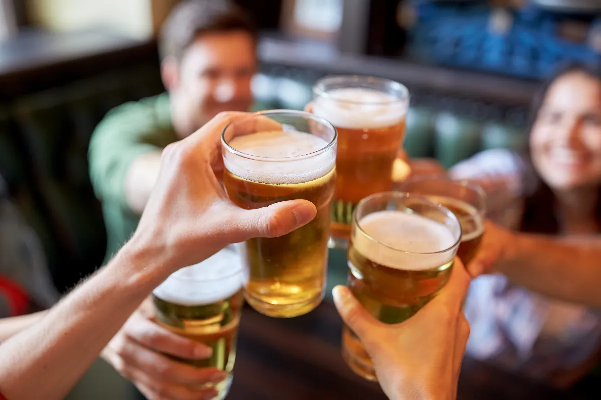 Grupo de amigos brindando com copos de cerveja em ambiente de bar, representando a comemoração do Dia da Cerveja com promoções e combos em estabelecimentos.