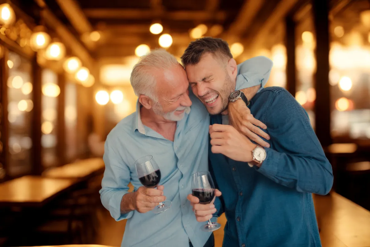 Pai e filho brindando com taças de vinho e sorrindo em restaurante aconchegante, representando o clima afetivo do Dia dos Pais e oportunidades de cardápios especiais.

