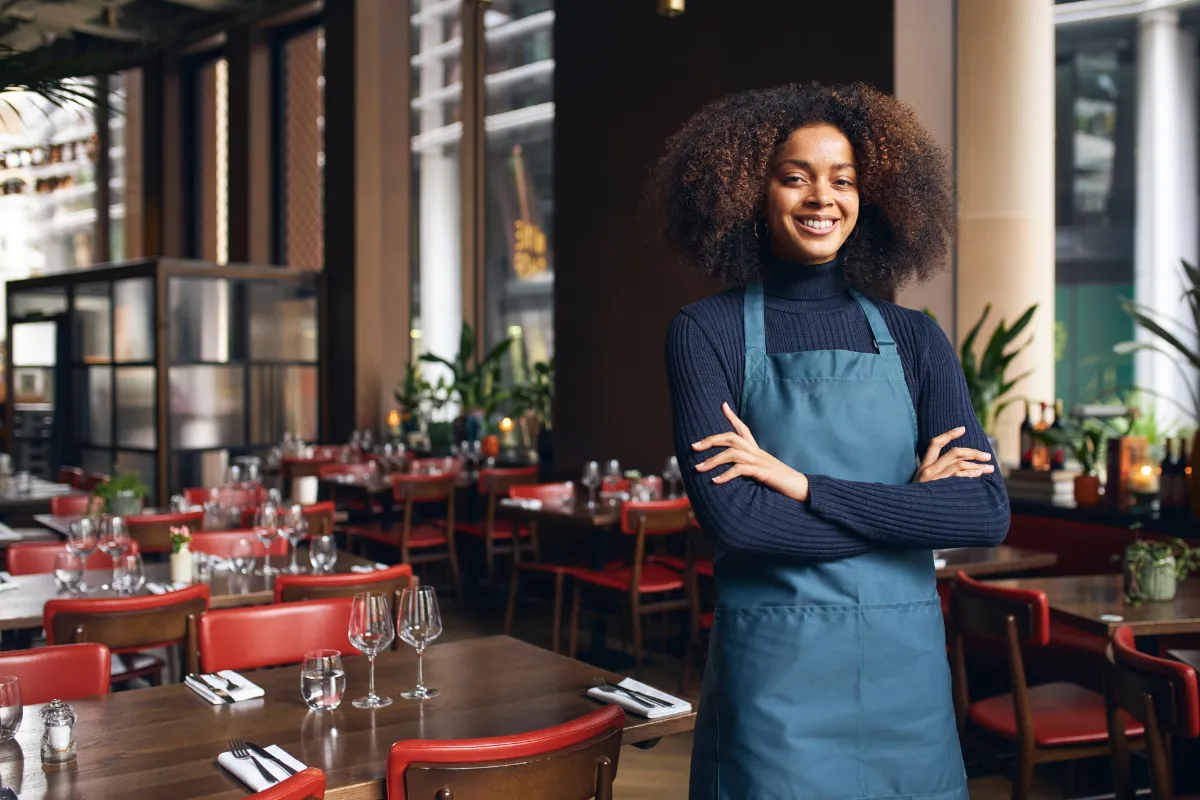 Empreendedora sorridente de avental azul em um restaurante arrumado e elegante, simbolizando o Dia Nacional da Mulher Empreendedora no setor de food service.