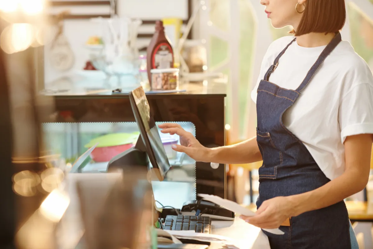 Colaboradora de cafeteria operando o sistema PDV e analisando comprovantes de pagamento durante o fechamento de caixa. A imagem ilustra a importância do registro detalhado das transações do dia.

