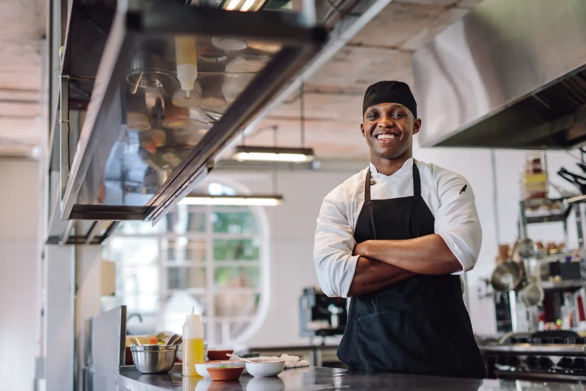 Chef de cozinha sorridente, com uniforme e avental preto, em uma cozinha profissional organizada — representando a importância do controle de estoque para manter a operação em pleno funcionamento no setor de Food Service.