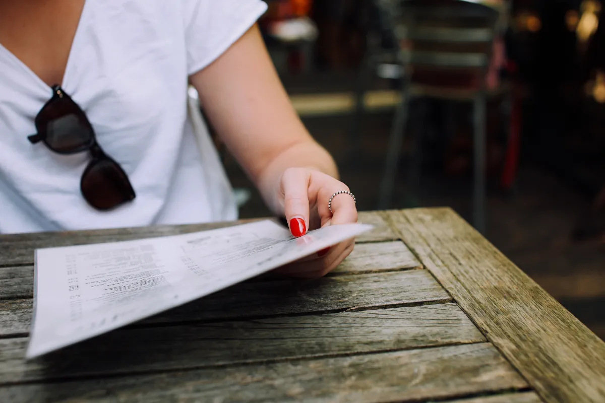 Cliente segurando cardápio impresso em papel, sentada em mesa de madeira ao ar livre, ilustrando a experiência tradicional de escolha.