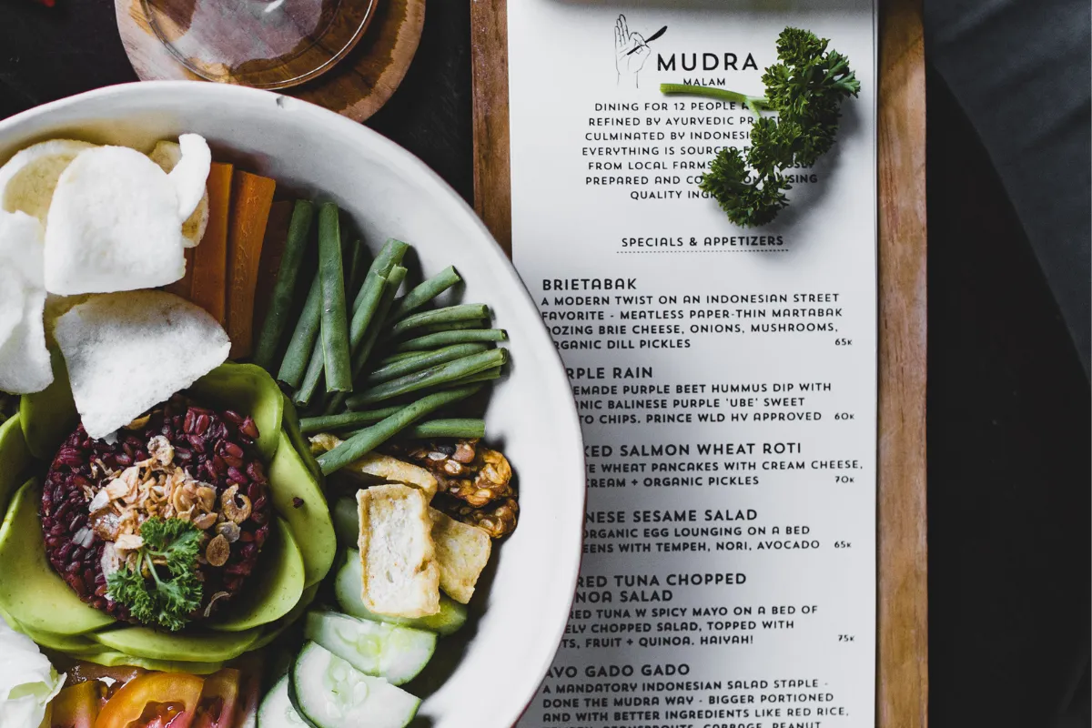 Cardápio impresso sofisticado ao lado de prato saudável com vegetais frescos, mostrando o design visual e gourmet do menu.