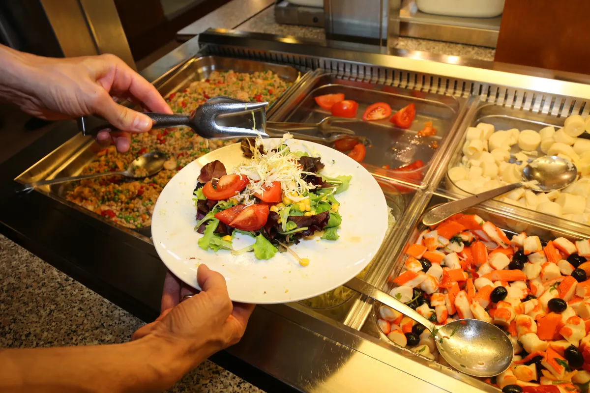 Pessoa montando prato com salada fresca em restaurante self-service, ilustrando a diversidade de opções e o controle necessário para reduzir desperdícios.